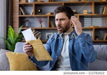 A surprised and concerned man sitting on a couch, holding and looking at a letter with a look of disbelief and worry. A surprised and concerned man sitting on a couch, holding and looking at a letter with a look of disbelief and worry. 112782622