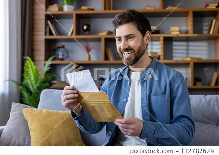 Happy adult male in a casual shirt, enjoying reading a letter while sitting comfortably on a sofa in a cozy living room. 112782629