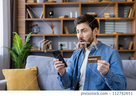 Man sitting on a couch looking shocked while holding a smartphone and a credit card, possibly after seeing unexpected charges. 112782651