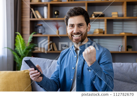 Joyful male sitting on a sofa with his fists pumped in triumph while holding a smartphone in a cozy living room setting. 112782670