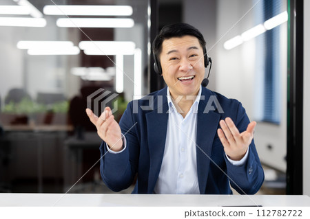 Professional smiling Asian businessman wearing a headset while engaged in an online video conference in a modern office setting. 112782722