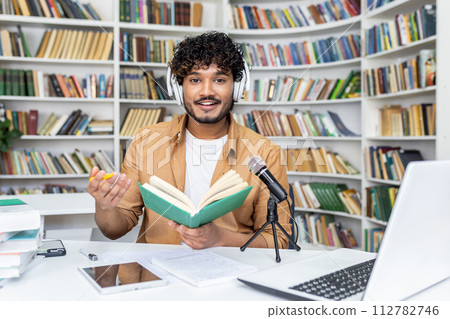 Confident blogger in white headset recording audio content while sitting with daybook by desk with professional micro. Islamic lecturer reading information for remote listeners in cyberspace. 112782746