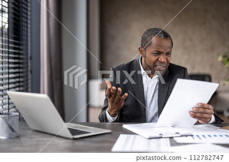 Confused male in shirt and jacket squinting in misunderstanding while reading information on paper shit. Overloaded worker getting unpleasantly surprised by amount of tasks while sitting by table. Confused male in shirt and jacket squinting in misunderstanding while reading information on paper shit. Overloaded worker getting unpleasantly surprised by amount of tasks while sitting by table. 112782747