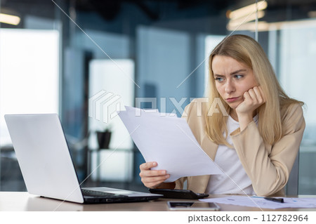 Focused young female in a business casual outfit sitting at her desk in a modern office setting, analyzing paperwork with a laptop beside her. 112782964