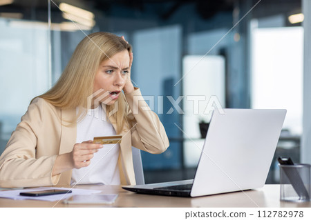 Businesswoman in office experiencing shock while holding a credit card and staring at a laptop screen, likely encountering unexpected charges. Businesswoman in office experiencing shock while holding a credit card and staring at a laptop screen, likely encountering unexpected charges. 112782978