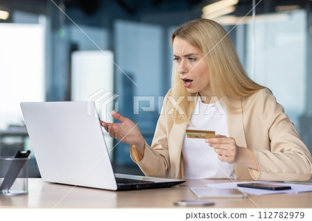 Shocked businesswoman holding a credit card looking at her laptop screen with confusion and surprise in an office setting. Shocked businesswoman holding a credit card looking at her laptop screen with confusion and surprise in an office setting. 112782979