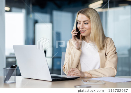 A businesswoman is engaged in conversation on her mobile phone while using a laptop in a modern office environment. 112782987