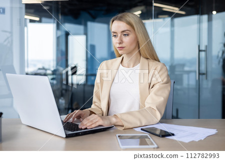 Professional woman in a beige blazer typing intently on a laptop at her workspace with smartphone and tablet in reach. Professional woman in a beige blazer typing intently on a laptop at her workspace with smartphone and tablet in reach. 112782993