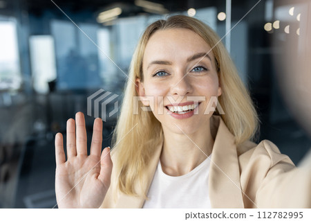 A cheerful young professional woman waving at the camera with a beaming smile in a contemporary office setting. A cheerful young professional woman waving at the camera with a beaming smile in a contemporary office setting. 112782995