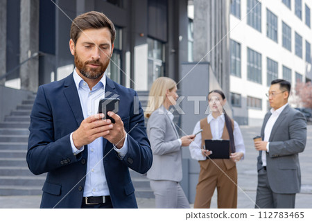 A group of corporate professionals engaged in conversation, checking smartphones during a work break outside modern office buildings. 112783465
