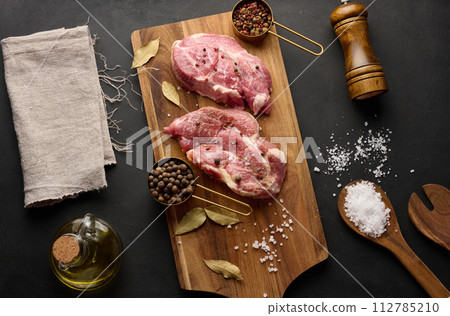 Two raw pork neck steaks on a board and spices for cooking. Top view of black table. Two raw pork neck steaks on a board and spices for cooking. Top view of black table. 112785210