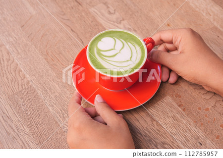 hand holding a matcha latte in a red color cup on table hand holding a matcha latte in a red color cup on table 112785957