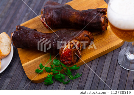 Picioare de porc afumate - juicy smoked pig legs on wooden board decorated with herbs, served with glass of cold beer and fresh bread Picioare de porc afumate - juicy smoked pig legs on wooden board decorated with herbs, served with glass of cold beer and fresh bread 112788519