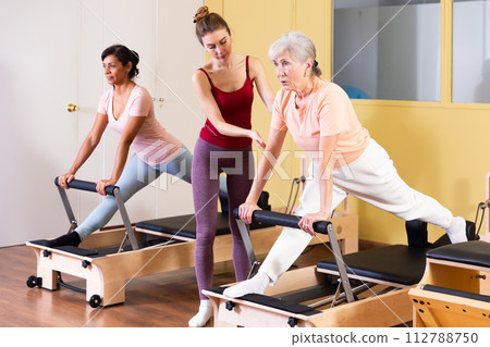 Young pilates instructor assisting elderly woman to do exercises on reformer 112788750