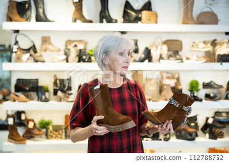 Contemplative elderly woman choosing demi-season boots in shoe store Contemplative elderly woman choosing demi-season boots in shoe store 112788752