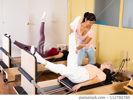 Aged woman doing pilates on reformer with Hispanic female trainer 112788780