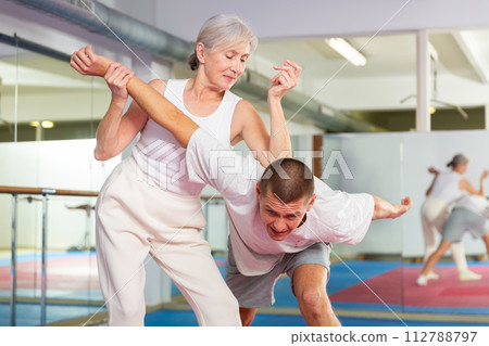 Elderly woman performing elbow strike and wristlock to man in self defense training Elderly woman performing elbow strike and wristlock to man in self defense training 112788797