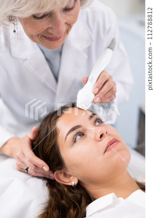 Professional mature female cosmetologist doing facial skin ultrasound cavitation for flawless woman face laying in beauty salon 112788812