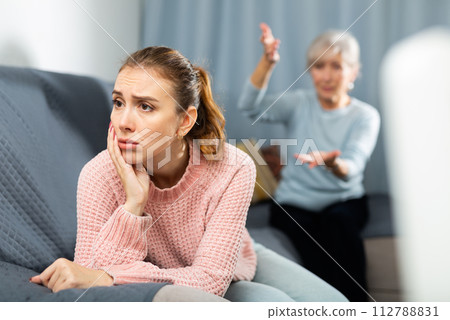 Upset young woman listening to reproaches from mother on sofa Upset young woman listening to reproaches from mother on sofa 112788831
