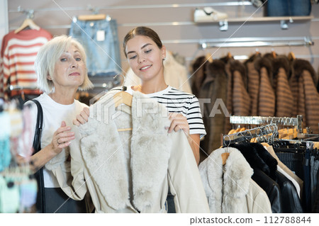 Girl consults with senior female friend about quality fur coat during shopping in clothes store Girl consults with senior female friend about quality fur coat during shopping in clothes store 112788844