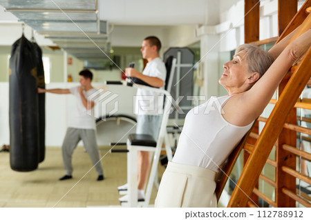 Elderly woman doing back stretching exercise on wall bar in gym 112788912