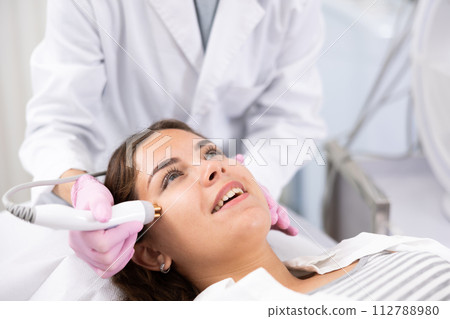 Mature female dermatologist performing cosmetology procedure for young woman, RF lifting and vacuum massage for facial skin therapy in clinic 112788980