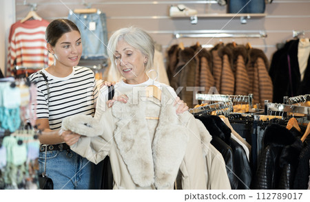 Girl consults with senior female friend about quality fur coat during shopping in clothes store Girl consults with senior female friend about quality fur coat during shopping in clothes store 112789017