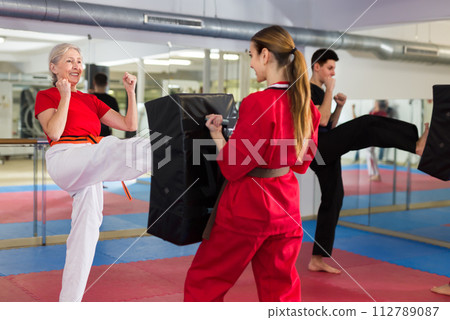 Aged woman kicking shield in martial arts training at gym Aged woman kicking shield in martial arts training at gym 112789087