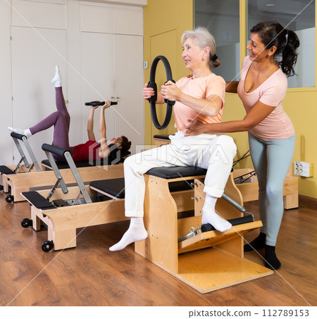 Elderly woman with trainer practicing pilates on combo chair with magic circle Elderly woman with trainer practicing pilates on combo chair with magic circle 112789153