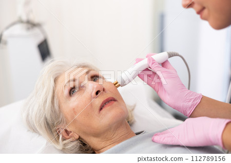 Young female dermatologist performing cosmetology procedure for old woman, RF lifting and vacuum massage for facial skin therapy in clinic 112789156