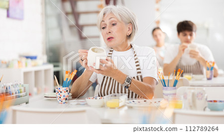 Talented senior female artisan applying design to ceramic mug in pottery studio Talented senior female artisan applying design to ceramic mug in pottery studio 112789264