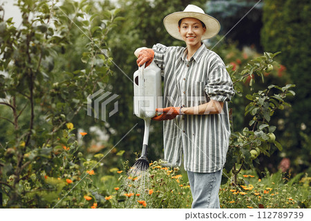 Gardening in summer. Woman watering flowers with a watering can. Girl wearing a hat. 112789739