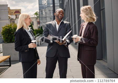 Confident businesspeople standing in front of modern office building. Black man and caucasian women talking and looking at documents. Banking and financial market concept. Confident businesspeople standing in front of modern office building. Black man and caucasian women talking and looking at documents. Banking and financial market concept. 112789764