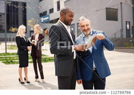 Close up on caucasian man wearing blue jacket and talking with black man. Two blurred businesswomen standing in front of modern office building. Banking and financial market concept. Close up on caucasian man wearing blue jacket and talking with black man. Two blurred businesswomen standing in front of modern office building. Banking and financial market concept. 112789766