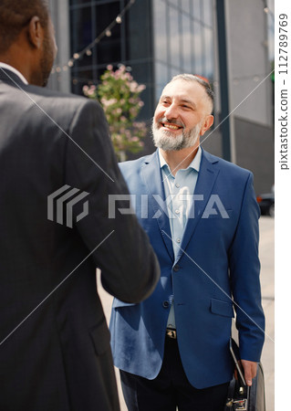Confident businesspeople standing in front of modern office building. Black and caucasian men shaking hands. Focus on a caucasian man. Confident businesspeople standing in front of modern office building. Black and caucasian men shaking hands. Focus on a caucasian man. 112789769
