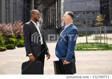 Confident businesspeople standing in front of modern office building. Black and caucasian men talking. Banking and financial market concept. Confident businesspeople standing in front of modern office building. Black and caucasian men talking. Banking and financial market concept. 112789771