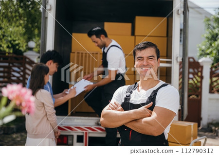 Portrait of a smiling mover unloading boxes into a new home from a truck. These removal company workers guarantee a joyful relocation and efficient service. Moving day concept Portrait of a smiling mover unloading boxes into a new home from a truck. These removal company workers guarantee a joyful relocation and efficient service. Moving day concept 112790312
