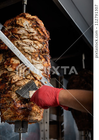Roasted meat on the bone for the preparation of donors or shawarma. Close-up. The cook cuts the meat fried over charcoal for the preparation of shawarma in pita bread.Close-up Roasted meat on the bone for the preparation of donors or shawarma. Close-up. The cook cuts the meat fried over charcoal for the preparation of shawarma in pita bread.Close-up 112791307