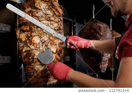 Roasted meat on the bone for the preparation of donors or shawarma. Close-up. The cook cuts the meat fried over charcoal for the preparation of shawarma in pita bread.Close-up Roasted meat on the bone for the preparation of donors or shawarma. Close-up. The cook cuts the meat fried over charcoal for the preparation of shawarma in pita bread.Close-up 112791314