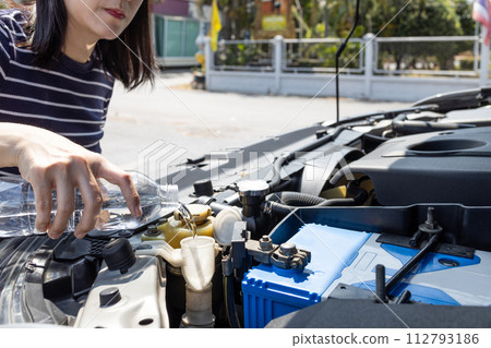 Asian woman pouring clean water from bottle into the windshield washer fluid tank of a car,female filling the windshield washer fluid,check liquid level in reservoir at engine room,maintenance concept 112793186