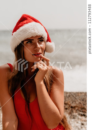 Woman summer travel sea. Happy tourist in red bikini and Santas hat enjoy taking picture outdoors for memories. Woman traveler posing on the beach surrounded by volcanic mountains, sharing travel joy 112793488