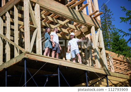 Man architect showcases to couple process of constructing wooden framed house. Youthful pair of investors inspecting their future dwelling in mountains. Concept of contemporary ecological construction Man architect showcases to couple process of constructing wooden framed house. Youthful pair of investors inspecting their future dwelling in mountains. Concept of contemporary ecological construction 112793909