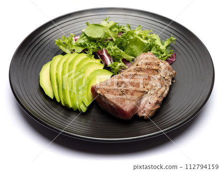 grilled cooked piece of tuna fillet with sliced avocado and salad on black ceramic plate 112794159