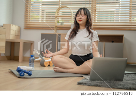 A calm, relaxed young Asian woman in sportswear is meditating on a yoga mat in the living room. 112796513