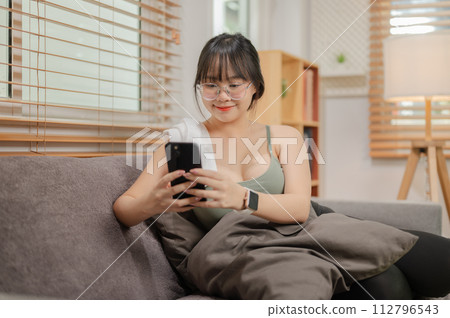 A woman is using her phone and relaxing on the sofa in her living room after finishing her workout. A woman is using her phone and relaxing on the sofa in her living room after finishing her workout. 112796543