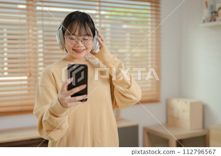 A cute Asian woman is listening to music on her headphones and using her phone in the living room. A cute Asian woman is listening to music on her headphones and using her phone in the living room. 112796567