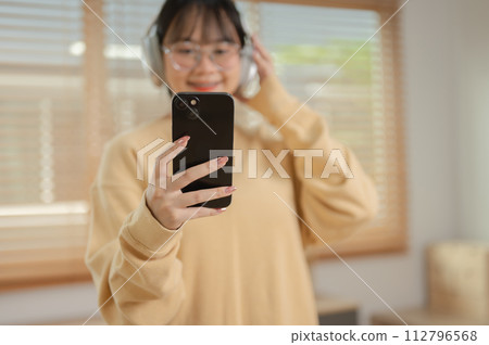 A cute Asian woman is listening to music on her headphones and using her phone in the living room. A cute Asian woman is listening to music on her headphones and using her phone in the living room. 112796568