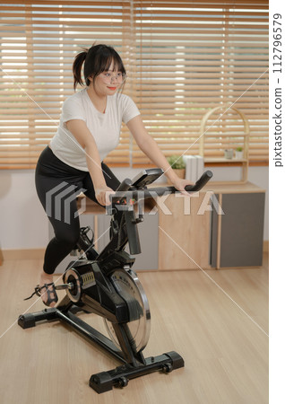 A strong Asian woman in sportswear is exercising on a fitness bike, doing indoor cardio at home. A strong Asian woman in sportswear is exercising on a fitness bike, doing indoor cardio at home. 112796579