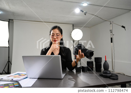 Creative young woman holding her DSLR camera and working with laptop at photo studio interior 112798474