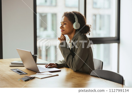 A woman at a table, using laptop with headphones, near a building window in nice modern coworking office 112799134